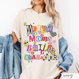 May include: Ivory-colored t-shirt with the text "WEIRD MOMS BUILD CHARACTER" in a colorful, collage-style design. The graphic includes flowers, stars, a checkered heart, and a melting skull. The shirt is a casual, oversized fit.