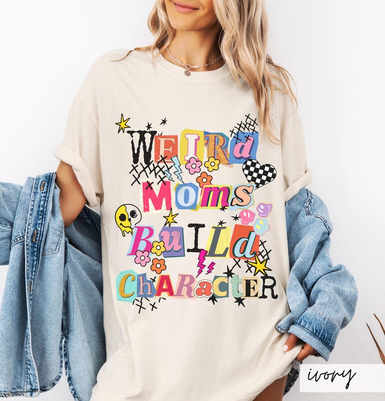 May include: Ivory-colored t-shirt with the text "WEIRD MOMS BUILD CHARACTER" in a colorful, collage-style design. The graphic includes flowers, stars, a checkered heart, and a melting skull. The shirt is a casual, oversized fit.