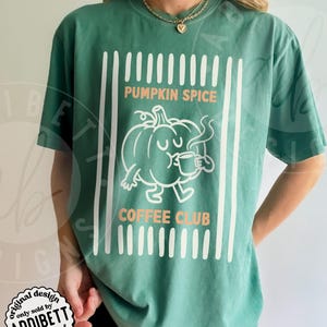 May include: A light green short-sleeve t-shirt featuring a white graphic of a cartoon pumpkin drinking from a mug. The text PUMPKIN SPICE COFFEE CLUB is printed in orange. The design is framed by vertical white stripes on the left and right sides.