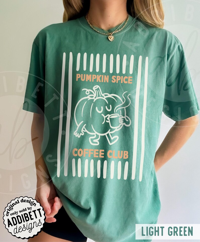 May include: A light green short-sleeve t-shirt featuring a white graphic of a cartoon pumpkin drinking from a mug. The text PUMPKIN SPICE COFFEE CLUB is printed in orange. The design is framed by vertical white stripes on the left and right sides.