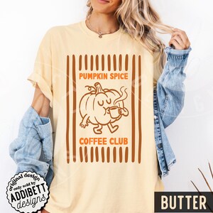 May include: A butter-coloured t-shirt featuring a brown graphic of a cartoon pumpkin drinking from a mug. The text reads PUMPKIN SPICE COFFEE CLUB. The design is framed by vertical brown stripes. The shirt is worn with a blue denim jacket.