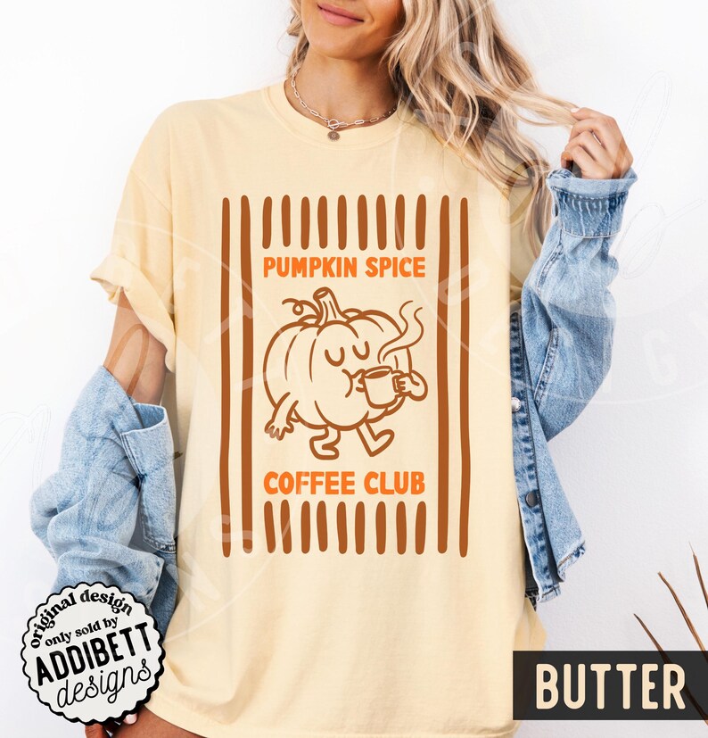 May include: A butter-coloured t-shirt featuring a brown graphic of a cartoon pumpkin drinking from a mug. The text reads PUMPKIN SPICE COFFEE CLUB. The design is framed by vertical brown stripes. The shirt is worn with a blue denim jacket.