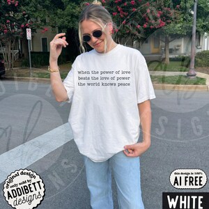 May include: A white short-sleeved t-shirt featuring black text on the chest that reads, when the power of love beats the love of power the world knows peace. The shirt is paired with light blue denim jeans and dark-tinted sunglasses.