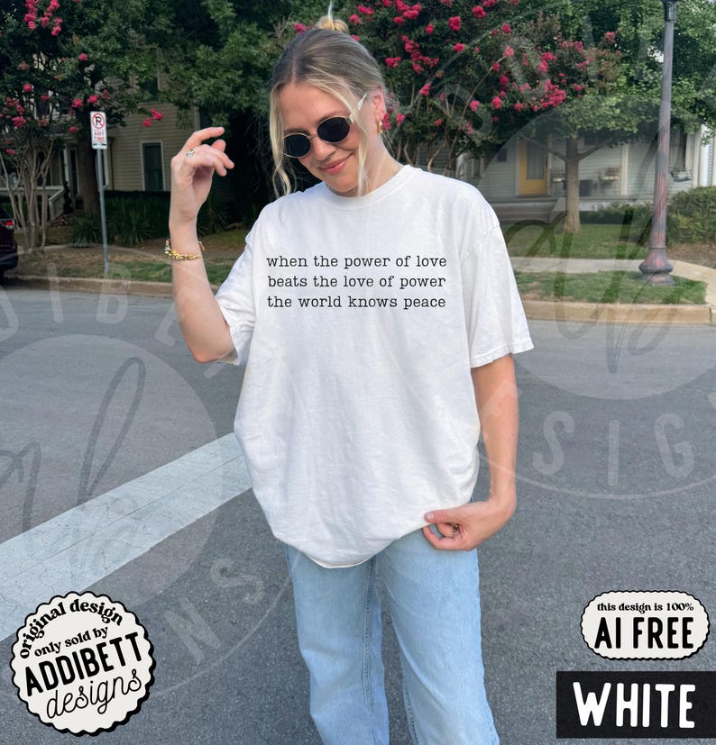 May include: A white short-sleeved t-shirt featuring black text on the chest that reads, when the power of love beats the love of power the world knows peace. The shirt is paired with light blue denim jeans and dark-tinted sunglasses.