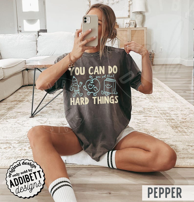 K&ouml;nnte beinhalten: Dunkelgraues T-Shirt mit dem Text "YOU CAN DO HARD THINGS" und einer Bleistift-, Apfel- und Buchgrafik. Das Shirt hat auch den Text "you got this!". Das Shirt ist Teil der "ADDIBETT designs" Kollektion.