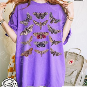 May include: A purple short-sleeve t-shirt featuring a graphic print of twelve different moth species in various shades of brown, gray, and red. The moths are arranged in a grid-like pattern across the front of the garment, showcasing detailed wing markings and textures.