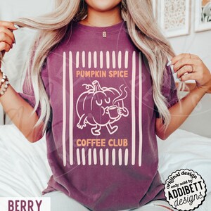 May include: A berry-coloured short-sleeve t-shirt featuring a graphic of a cartoon pumpkin holding a coffee mug. The text reads PUMPKIN SPICE COFFEE CLUB. The design is framed by vertical stripes in a light cream colour on a solid berry background.