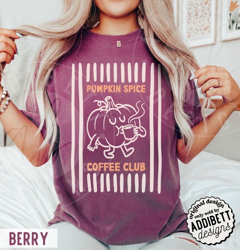 May include: A berry-coloured short-sleeve t-shirt featuring a graphic of a cartoon pumpkin holding a coffee mug. The text reads PUMPKIN SPICE COFFEE CLUB. The design is framed by vertical stripes in a light cream colour on a solid berry background.