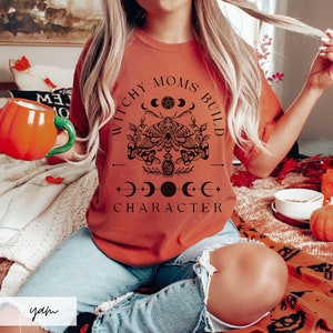 May include: A woman wearing a burnt orange t-shirt with the text "Witchy Moms Build Character" in black lettering. The design includes a moth, crescent moons, and a potion bottle.