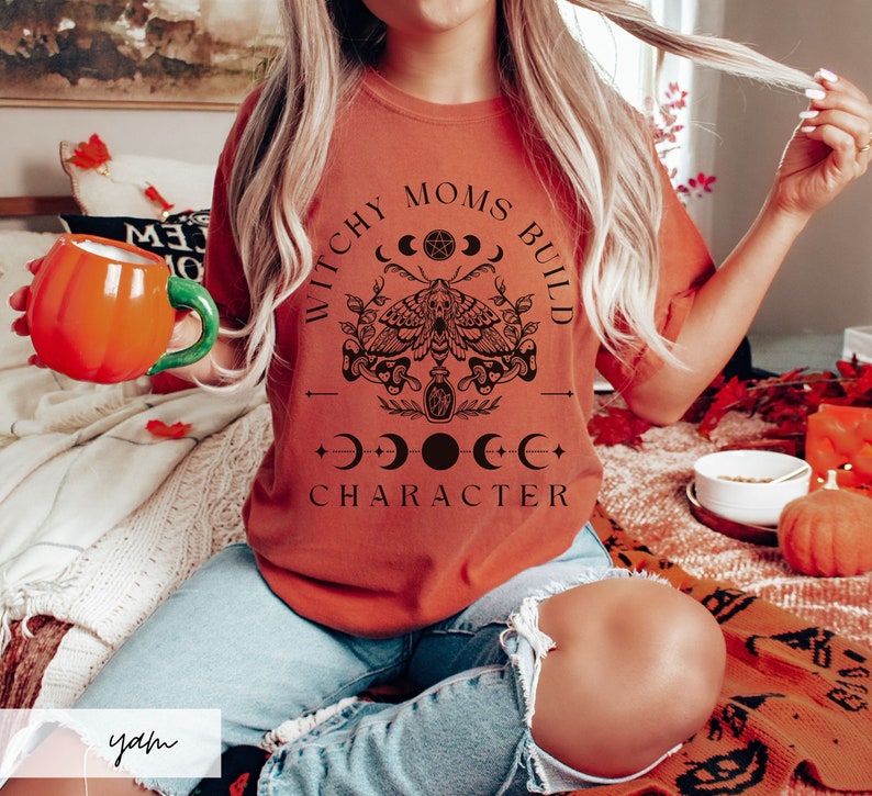 May include: A woman wearing a burnt orange t-shirt with the text "Witchy Moms Build Character" in black lettering. The design includes a moth, crescent moons, and a potion bottle.