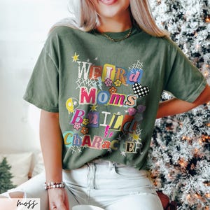 May include: Moss green t-shirt with the text "WEIRD Moms BUILD CHARACTER" in a colorful, collage-style design. The shirt features a crew neck and short sleeves. The text includes flowers, stars, and a checkered heart.