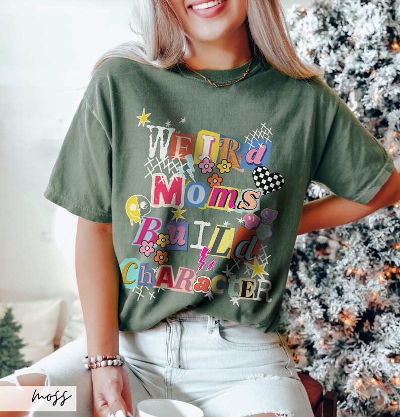 May include: Moss green t-shirt with the text "WEIRD Moms BUILD CHARACTER" in a colorful, collage-style design. The shirt features a crew neck and short sleeves. The text includes flowers, stars, and a checkered heart.