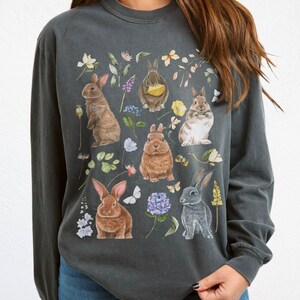 May include: A charcoal grey long-sleeved t-shirt with a graphic print of multiple rabbits and flowers. The rabbits are brown, grey, and white.