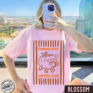 May include: A light pink t-shirt featuring a graphic print of a cartoon pumpkin holding a small mug. The text PUMPKIN SPICE COFFEE CLUB is printed in orange above and below the pumpkin, framed by vertical orange stripes on the sides.