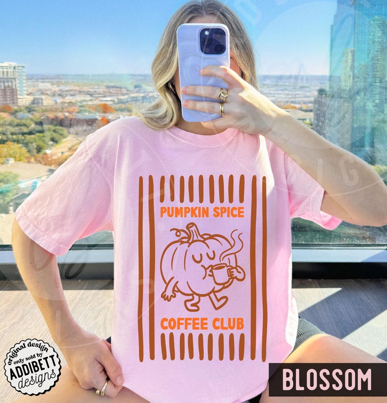 May include: A light pink t-shirt featuring a graphic print of a cartoon pumpkin holding a small mug. The text PUMPKIN SPICE COFFEE CLUB is printed in orange above and below the pumpkin, framed by vertical orange stripes on the sides.