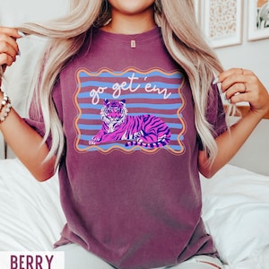 Pu&ograve; includere: T-shirt color bacca con un'illustrazione di tigre e la scritta "go get 'em". La tigre &egrave; raffigurata in rosa e viola con strisce nere, su uno sfondo a righe blu e arancioni.