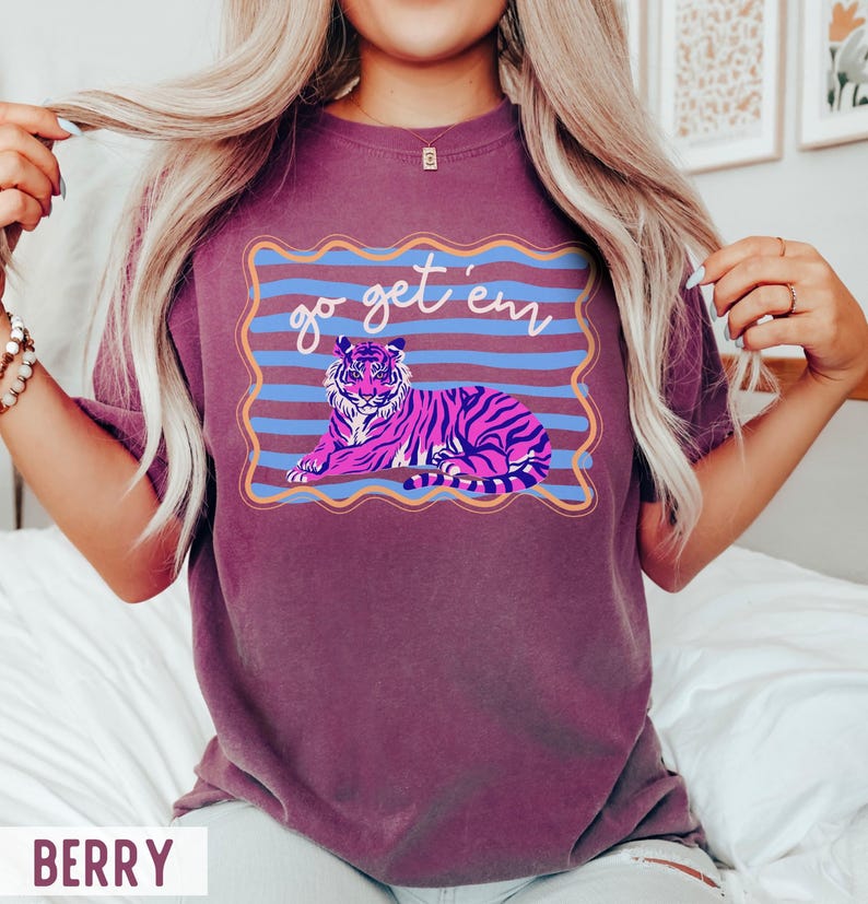 Pu&ograve; includere: T-shirt color bacca con un'illustrazione di tigre e la scritta "go get 'em". La tigre &egrave; raffigurata in rosa e viola con strisce nere, su uno sfondo a righe blu e arancioni.