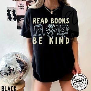 May include: A black short-sleeved t-shirt featuring the text READ BOOKS BE KIND in a light yellow font. Between the lines of text are three white line-art illustrations of a smiling book, a smiling flower pot, and an open smiling book.