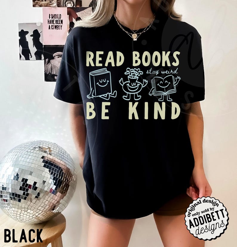 May include: A black short-sleeved t-shirt featuring the text READ BOOKS BE KIND in a light yellow font. Between the lines of text are three white line-art illustrations of a smiling book, a smiling flower pot, and an open smiling book.