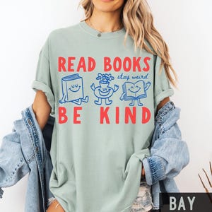 May include: A sage green short-sleeve t-shirt featuring the red text READ BOOKS BE KIND and blue line art illustrations of a smiling book, a smiling flower vase, and an open smiling book with the blue script text stay weird in the center.