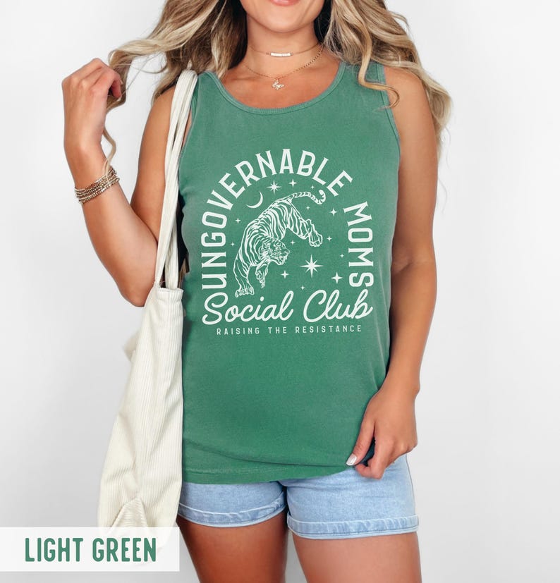 Puede incluir: Camiseta sin mangas verde claro con el texto "YOUNG GOVERNABLE MOMS SOCIAL CLUB RAISING THE RESISTANCE" y un gr&aacute;fico de tigre. La camiseta sin mangas se combina con pantalones cortos de mezclilla azules.