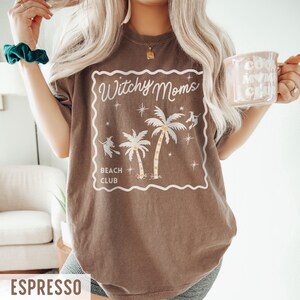 K&ouml;nnte beinhalten: Espressofarbenes T-Shirt mit wei&szlig;em Aufdruck &bdquo;Witchy Moms Beach Club&ldquo; mit Palmen und Hexensilhouetten. Das Shirt hat eine lockere Passform und kurze &Auml;rmel. Eine hellrosa Tasse mit wei&szlig;em Text wird in der Hand gehalten.