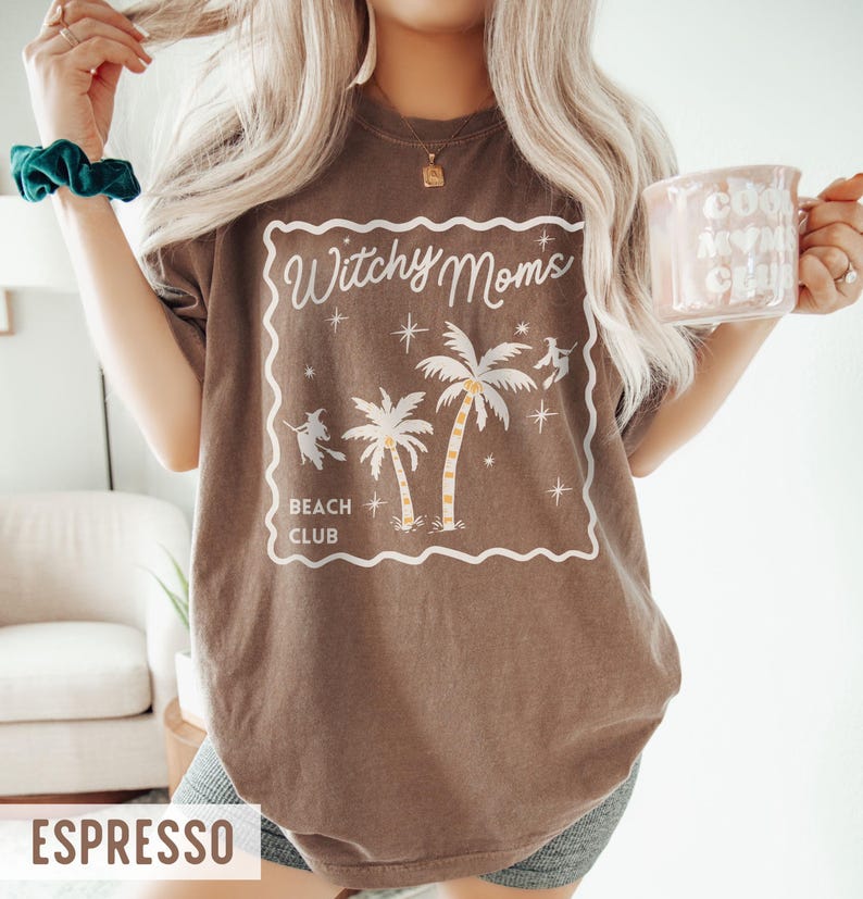 K&ouml;nnte beinhalten: Espressofarbenes T-Shirt mit wei&szlig;em Aufdruck &bdquo;Witchy Moms Beach Club&ldquo; mit Palmen und Hexensilhouetten. Das Shirt hat eine lockere Passform und kurze &Auml;rmel. Eine hellrosa Tasse mit wei&szlig;em Text wird in der Hand gehalten.