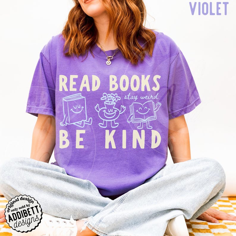 May include: A violet short-sleeved t-shirt featuring the white text READ BOOKS BE KIND and stay weird. The graphic includes three smiling cartoon characters: a closed book, a flower in a vase, and an open book, all with arms and legs.