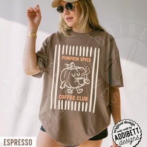 May include: A brown short-sleeve t-shirt featuring a graphic of a cartoon pumpkin holding a mug. The text reads PUMPKIN SPICE COFFEE CLUB in light orange letters. The design is framed by vertical white lines on a brown fabric background.