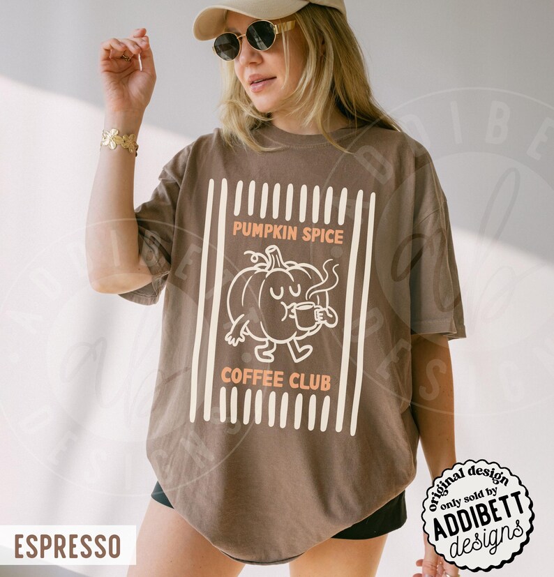 May include: A brown short-sleeve t-shirt featuring a graphic of a cartoon pumpkin holding a mug. The text reads PUMPKIN SPICE COFFEE CLUB in light orange letters. The design is framed by vertical white lines on a brown fabric background.