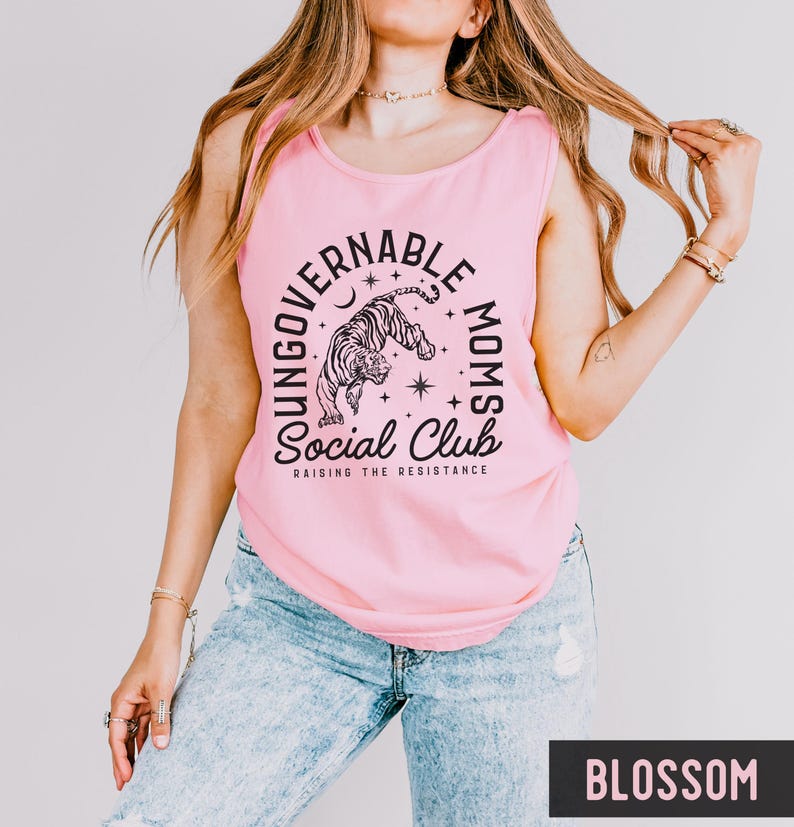 Puede incluir: Una camiseta sin mangas rosa claro con el texto "YOUNGGOVERNABLE MOMS Social Club RAISING THE RESISTANCE" y un gr&aacute;fico de tigre. La camiseta sin mangas se combina con vaqueros azul claro. La palabra "BLOSSOM" est&aacute; en un recuadro negro.