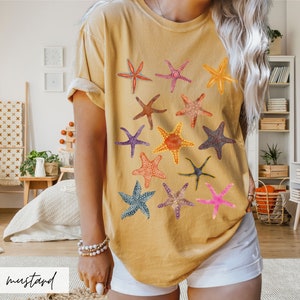May include: Mustard-colored t-shirt featuring a collection of colorful starfish designs. The starfish vary in color and pattern, including red, orange, purple, and blue. The word "mustard" is written in a cursive font at the bottom of the image.