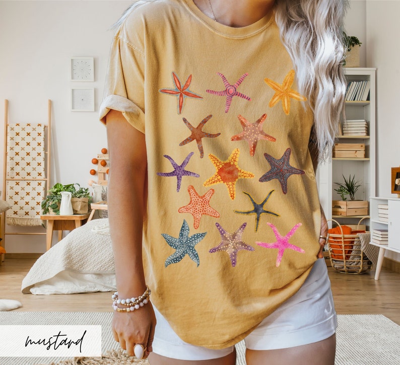 May include: Mustard-colored t-shirt featuring a collection of colorful starfish designs. The starfish vary in color and pattern, including red, orange, purple, and blue. The word "mustard" is written in a cursive font at the bottom of the image.