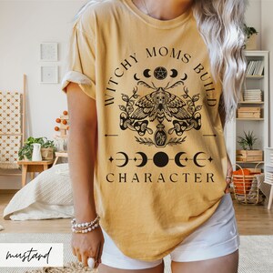 May include: A mustard yellow t-shirt with a black graphic design that says "Witchy Moms Build Character" with a moth, moons, and a pentagram.