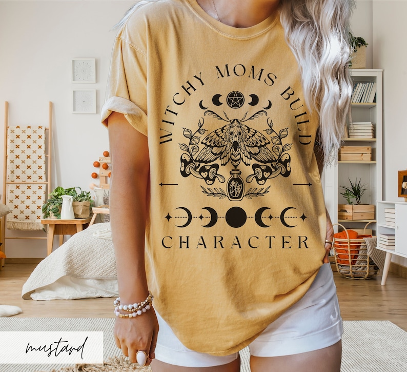 May include: A mustard yellow t-shirt with a black graphic design that says "Witchy Moms Build Character" with a moth, moons, and a pentagram.