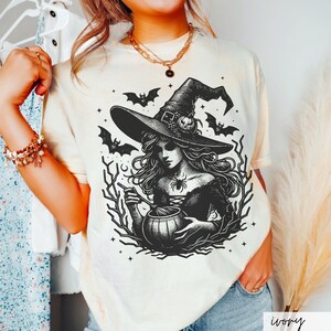 May include: A white t-shirt with a black and white illustration of a witch stirring a cauldron. The witch is wearing a pointed hat and has long, flowing hair. The illustration is surrounded by bats and stars.
