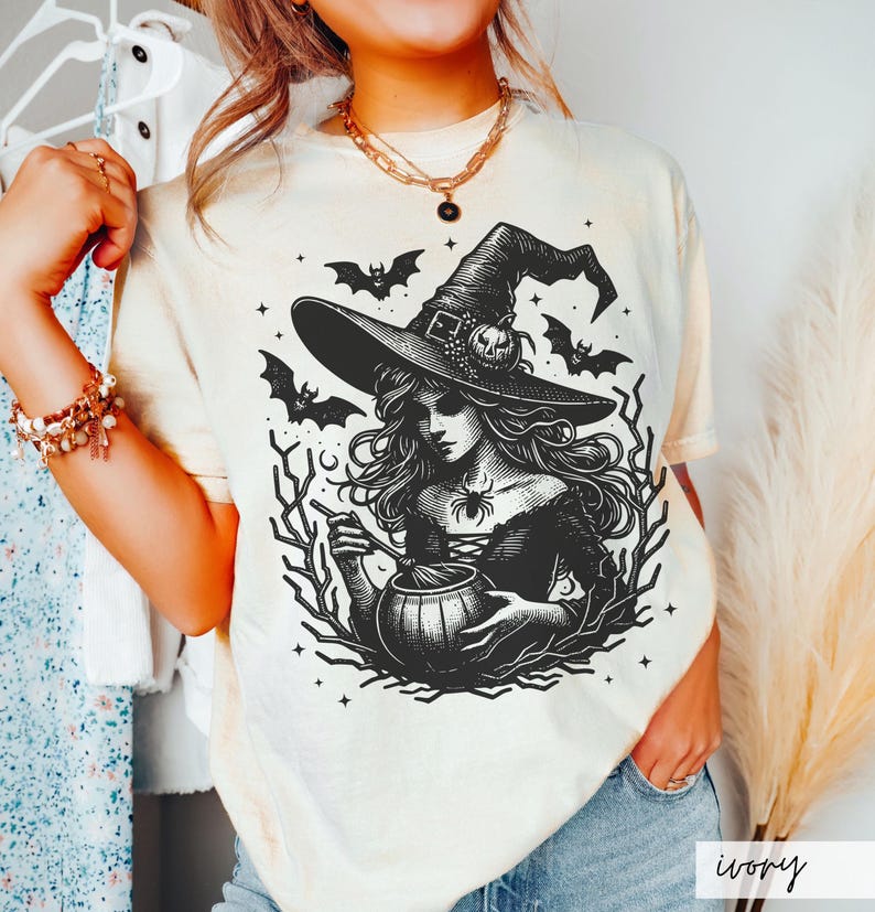 May include: A white t-shirt with a black and white illustration of a witch stirring a cauldron. The witch is wearing a pointed hat and has long, flowing hair. The illustration is surrounded by bats and stars.