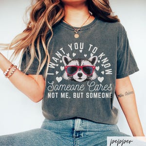 以下が含まれることがあります： 赤いサングラスをかけたアライグマと「I want you to know someone cares not me, but someone」というテキストがプリントされたダークグレーのTシャツです。