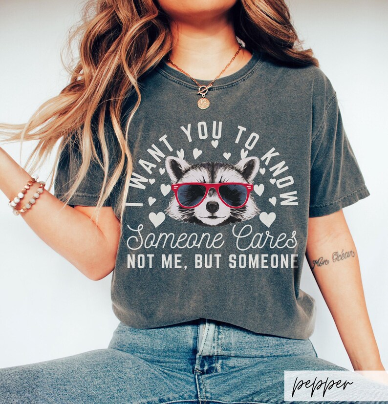 以下が含まれることがあります： 赤いサングラスをかけたアライグマと「I want you to know someone cares not me, but someone」というテキストがプリントされたダークグレーのTシャツです。