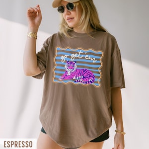 Pu&ograve; includere: T-shirt color espresso con grafica di una tigre rosa e viola e la scritta "go get 'em" in corsivo bianco. La tigre &egrave; all'interno di un bordo a righe blu e marroni. La maglietta ha una vestibilit&agrave; ampia.