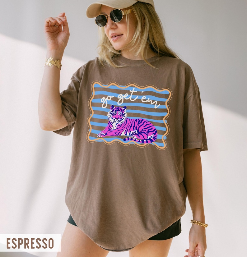 Pu&ograve; includere: T-shirt color espresso con grafica di una tigre rosa e viola e la scritta "go get 'em" in corsivo bianco. La tigre &egrave; all'interno di un bordo a righe blu e marroni. La maglietta ha una vestibilit&agrave; ampia.
