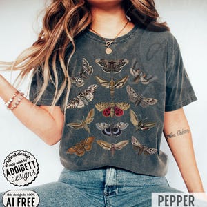 May include: A charcoal gray short-sleeve t-shirt featuring a graphic print of twelve different moth species in various shades of brown, tan, and red. The shirt has a crew neckline and a vintage-style washed finish across the fabric surface.