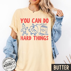 K&ouml;nnte beinhalten: Ein hellgelbes T-Shirt mit dem Text "YOU CAN DO HARD THINGS" in Rot. Das Design zeigt blaue Cartoon-Illustrationen eines Bleistifts, Apfels und Buches. Der Satz "you got this!" steht &uuml;ber dem Apfel. Das Wort "BUTTER" steht unten.