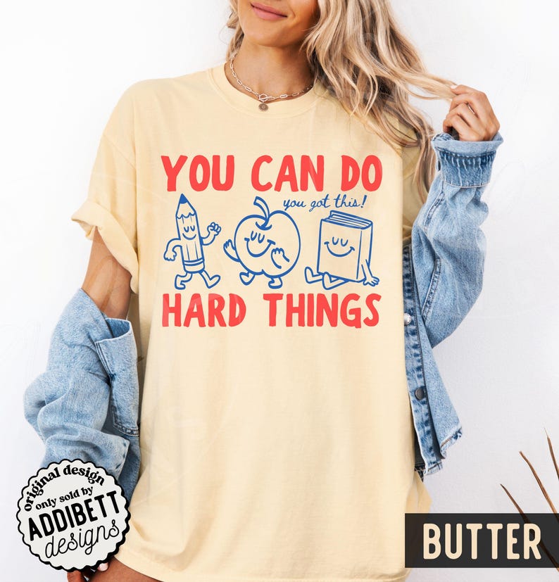 K&ouml;nnte beinhalten: Ein hellgelbes T-Shirt mit dem Text "YOU CAN DO HARD THINGS" in Rot. Das Design zeigt blaue Cartoon-Illustrationen eines Bleistifts, Apfels und Buches. Der Satz "you got this!" steht &uuml;ber dem Apfel. Das Wort "BUTTER" steht unten.