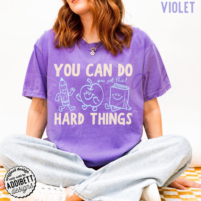 K&ouml;nnte beinhalten: Ein violettes T-Shirt mit dem Text "YOU CAN DO HARD THINGS" und dem Satz "you got this!". Das Design zeigt Cartoon-Illustrationen eines Bleistifts, eines Apfels und eines Buches. Das Shirt hat eine einfarbige violette Farbe.