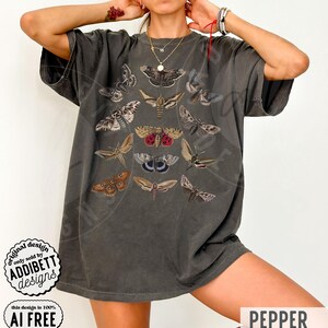 May include: A charcoal gray short-sleeve t-shirt featuring a central graphic print of various moth species in shades of brown, tan, and red. The shirt has a crew neckline and a loose, relaxed fit. A circular logo in the corner reads ADDIBETT DESIGNS.