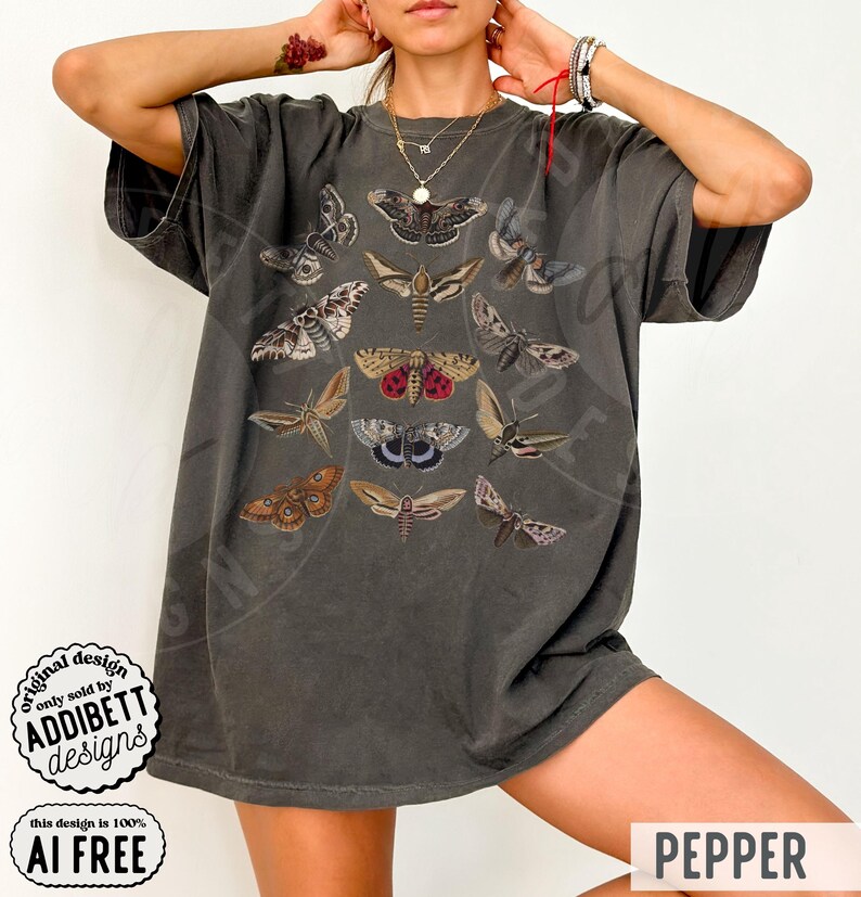 May include: A charcoal gray short-sleeve t-shirt featuring a central graphic print of various moth species in shades of brown, tan, and red. The shirt has a crew neckline and a loose, relaxed fit. A circular logo in the corner reads ADDIBETT DESIGNS.