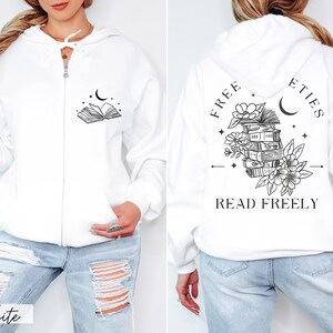 May include: White zip-up hoodie with a hood and front pockets. The front features a black graphic of an open book, a crescent moon, and stars. The back displays a stack of books, flowers, and the text "FREE ETIES READ FREELY".