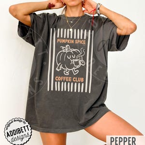 May include: A charcoal grey short-sleeve t-shirt featuring a white line art graphic of a pumpkin character holding a coffee mug. The text reads PUMPKIN SPICE COFFEE CLUB in light orange letters, framed by vertical white stripes on a dark background.