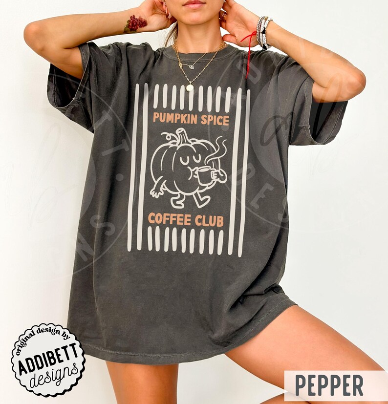 May include: A charcoal grey short-sleeve t-shirt featuring a white line art graphic of a pumpkin character holding a coffee mug. The text reads PUMPKIN SPICE COFFEE CLUB in light orange letters, framed by vertical white stripes on a dark background.